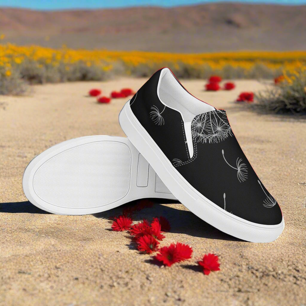 Black Dandelion Floral Print Slip-on Canvas Shoes on a sandy path with flowers in the background