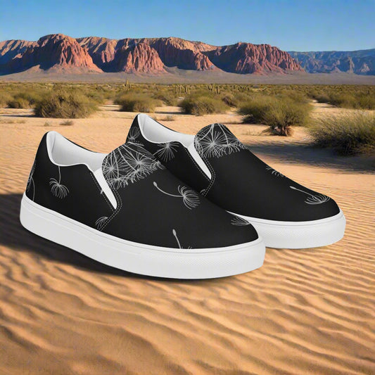 Black Dandelion Floral Print Slip-on Canvas Shoes on a sandy desert landscape with mountains in the background