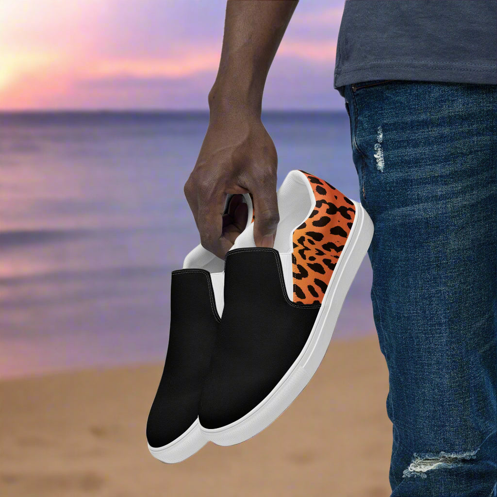 Mens Cheetah Two Tone Slip on Canvas Shoes with black front and cheetah print back held by a person near the beach