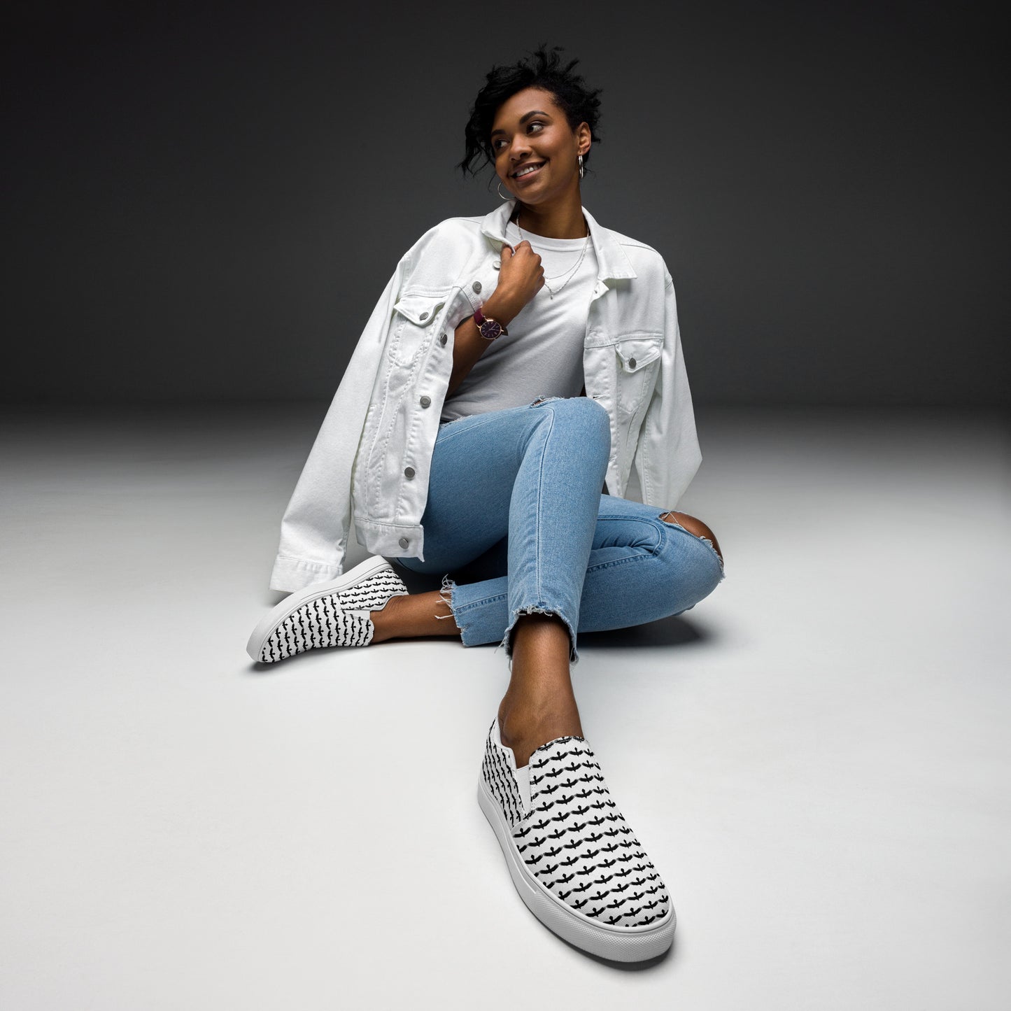 Black raven pattern slip on shoes worn by woman sitting wearing white jacket and blue jeans