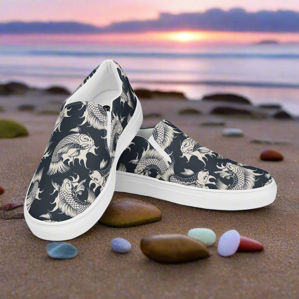 Black Koi Asian Print Slip-on Canvas Shoes resting on a beach with colorful stones